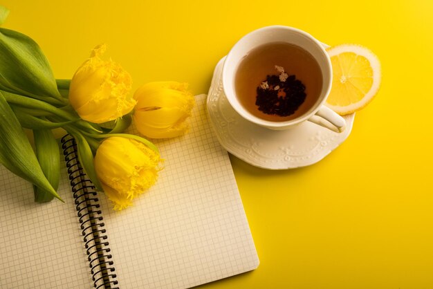 Fiori di tulipani gialli con tazza di tè al limone e blocco note con limone su sfondo giallo