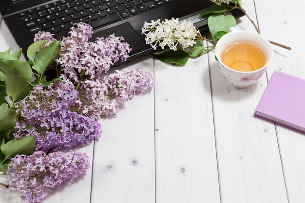 fiori lilla con taccuino viola aperto, laptop aperto nero, tazza da tè, vista prospettica mockup
