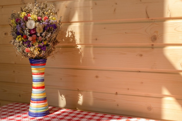 Fiori luminosi in vaso colorato contro la parete del pannello di legno sulla tovaglia a quadri rossa e bianca.