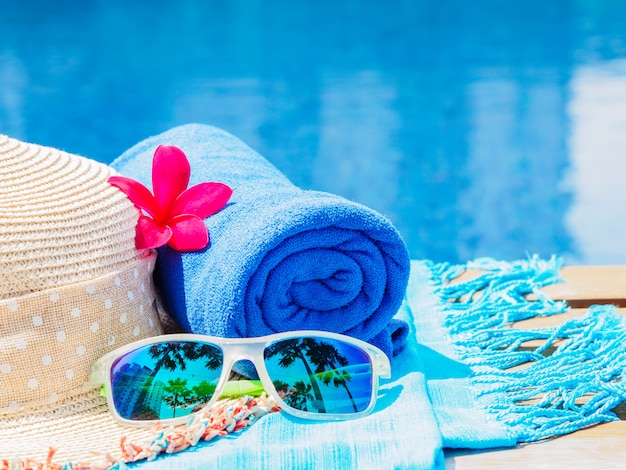 Fiori, occhiali da sole, cappello da spiaggia e asciugamano blu sul lato della piscina.