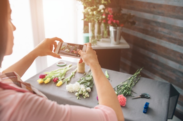 Fiorista femminile al lavoro: bella giovane donna dai capelli scuri che fa moda bouquet moderno di fiori diversi. Donne che lavorano con i fiori in officina. Sta fotografando fiori su uno smartphone.