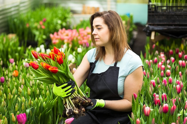 Fiorista giardiniere donna sorridente che tiene un mazzo di fiori in piedi in una serra dove coltivano i tulipani