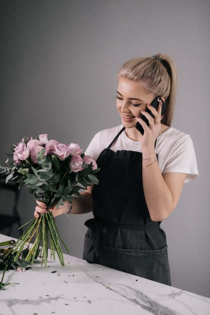 Fiorista professionista giovane donna che indossa il grembiule parlando al telefono cellulare con il cliente in possesso di meravigliosa composizione di piante fresche su sfondo bianco Concetto di lavorare con fiori affari floreali