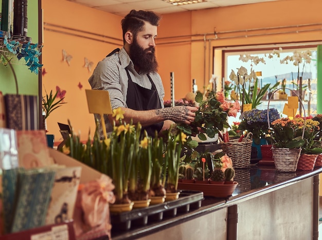 Fiorista professionista maschio con barba e tatuaggio sulla sua mano che fa una bella composizione floreale stando in piedi al bancone di un negozio di fiori.