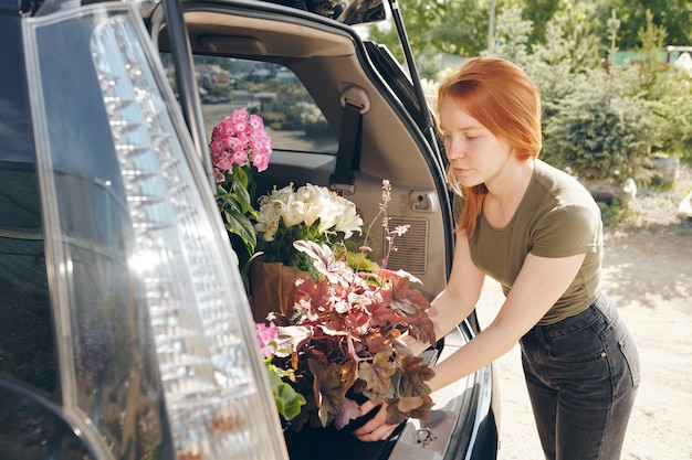 Fiorista rossa attraente serio in vestiti comodi che mette i fiori nel bagagliaio dell'auto mentre acquistano piante al mercato