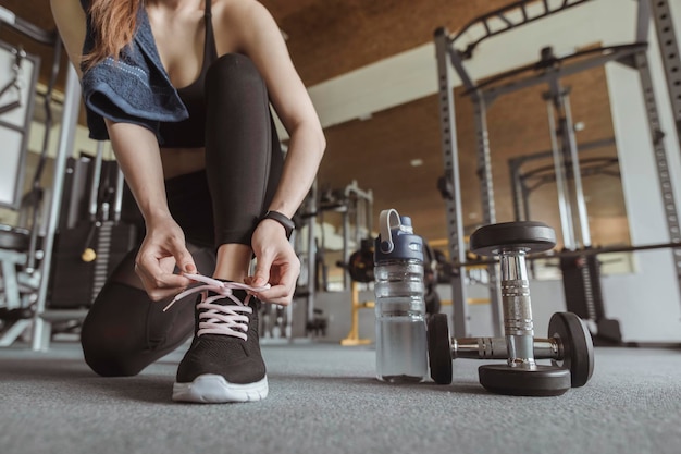 Fitness, allenamento, esercizio in palestra, stile di vita e concetto sano. Le donne usano le mani per allacciarsi le scarpe, hanno un manubrio e una bottiglia d'acqua accanto a loro in palestra per allenarsi al tramonto.