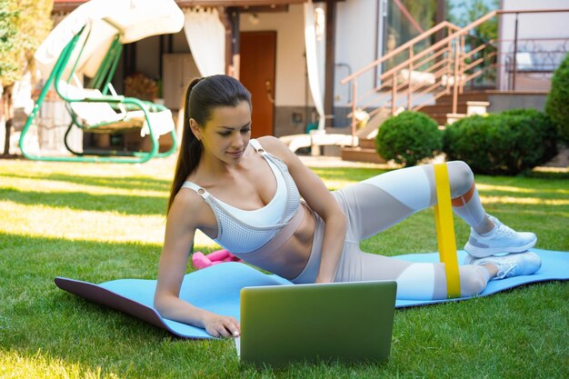 Fitness bella donna magra che fa tavola laterale con fascia di resistenza e guarda tutorial online sull'allenamento del laptop all'aperto