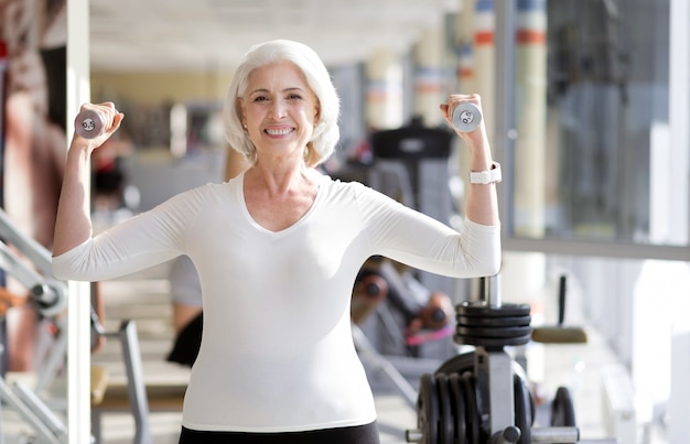 Fitness come stile di vita. Gioiosa sportiva donna senior mantenersi in forma e sorridente durante l'utilizzo di pesi durante l'esercizio in palestra.