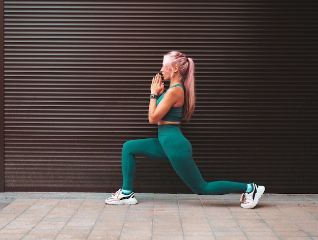 Fitness donna sorridente in abbigliamento sportivo grigio con capelli rosa Giovane bella modella con un corpo perfettoFemmina in posa per strada vicino al muroFare affondi in avanti Allungarsi prima dell'allenamento