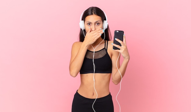 Fitness ispanica donna che copre la bocca con le mani con uno scioccato con le cuffie e un telefono