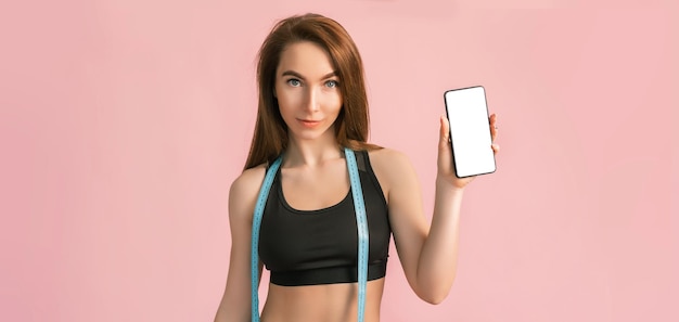 Fitness ragazza sorridente e in possesso di un telefono con un mockup e in posa tenere metro a nastro in abbigliamento sportivo nero su sfondo rosa Donna snella con un bel corpo atletico e pelle abbronzata