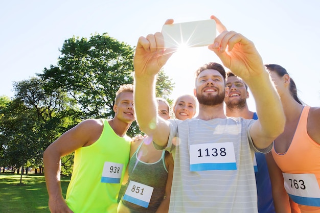 fitness, sport, amicizia, tecnologia e concetto di stile di vita sano - gruppo di amici sportivi felici con numeri di badge da corsa che prendono smartphone selfie all'aperto