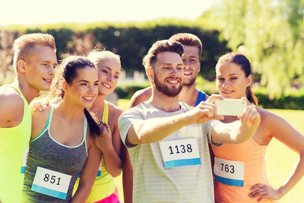 fitness, sport, amicizia, tecnologia e concetto di stile di vita sano - gruppo di amici sportivi felici con numeri di badge da corsa che prendono smartphone selfie all'aperto