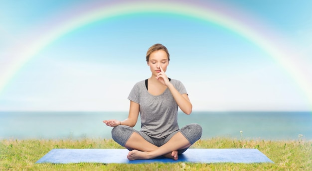 fitness, sport, persone e concetto di stile di vita sano - donna che fa meditazione yoga nella posa del loto sul tappeto sopra il cielo blu, l'arcobaleno e lo sfondo del mare