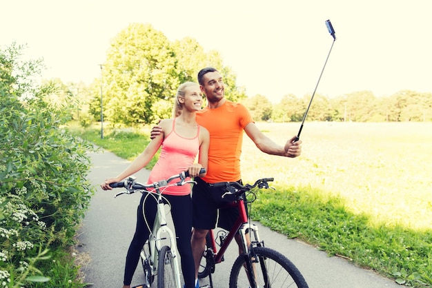 fitness, sport, persone, tecnologia e concetto di stile di vita sano - coppia felice con la bicicletta che scatta foto con lo smartphone sul selfie stick all'aperto