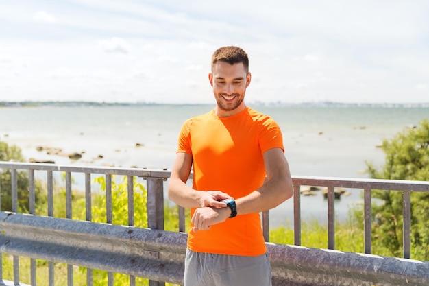 fitness, sport, persone, tecnologia e concetto di stile di vita sano - giovane sorridente con orologio da polso intelligente al mare estivo