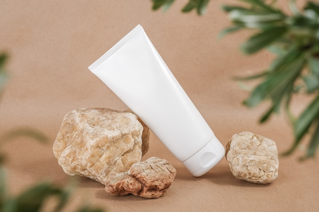 Flacone cosmetico vuoto bianco tubo su pietra incorniciato da foglie verdi di rami, sfondo beige Organico naturale