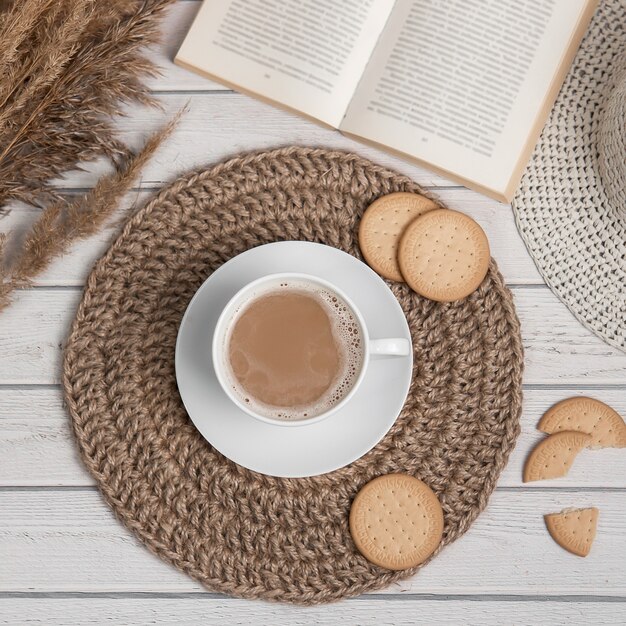 Flatlay con tazza di biscotti di stuoia di iuta americano o espresso e libro sul tavolo di legno bianco.