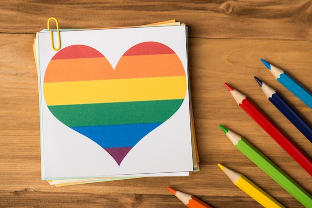 Flatlay foto ravvicinata di pile di adesivi di carta con cuore arcobaleno disegnato e matite multicolori isolato sullo sfondo di legno