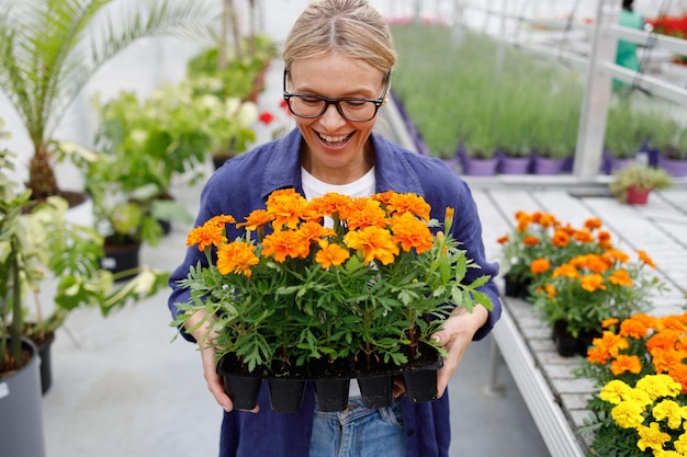 Florista donna gioiosa che tiene un vaso con fiori di marigold in serra