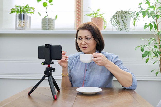 Flusso di vlog del blog video, donna che guarda la webcam dello smartphone sul treppiede, registra video, parla online, si siede a tavola a casa. Canale educativo, giornalista, psicologo, counselor