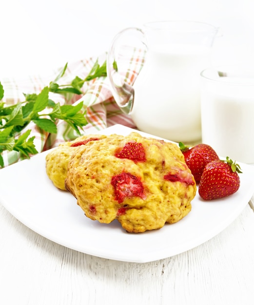 Focaccine alla fragola in un piatto con frutti di bosco, un tovagliolo, menta, latte in brocca e vetro sullo sfondo di una tavola di legno chiaro