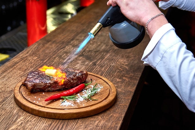 Focalizzazione morbida. Entrecote Manzo Bistecca alla griglia con fiamme di fuoco su tagliere di legno con ramo di rosmarino, pepe e sale. Master chef che cucina deliziosi barbecue alla griglia. Sciogliere il burro