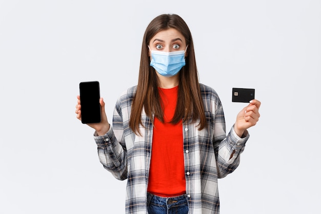 Focolaio di coronavirus, lavoro da casa, shopping online e concetto di pagamento senza contatto. Donna eccitata in maschera medica che mostra carta di credito e telefono cellulare, sembra sorpresa