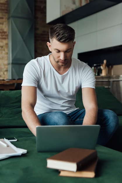 Focus selettivo del bel libero professionista che utilizza il laptop vicino a documenti e libri sul divano