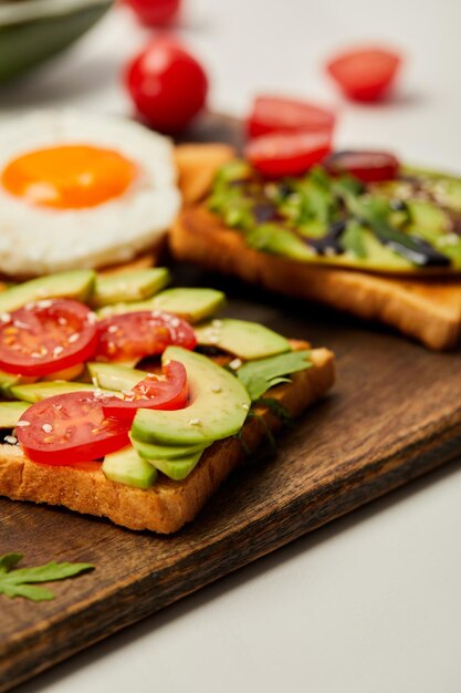 Focus selettivo del tagliere con toast uova strapazzate pomodorini e avocado su bianco