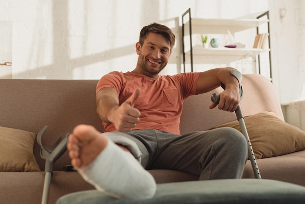 Focus selettivo dell'uomo sorridente con la gamba rotta che mostra il simbolo del pollice sul divano di casa