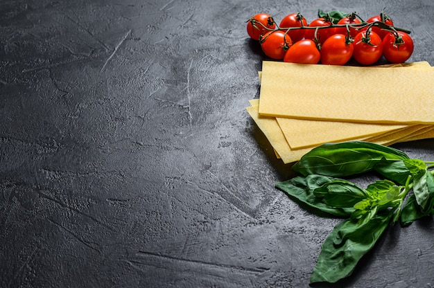 Fogli di lasagne crude. Ingredienti Basilico, pomodorini. Sfondo nero. Vista dall'alto. Spazio per il testo