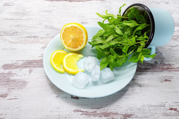 Foglie di menta in tazza, limone e cubetti di ghiaccio sulla zolla blu su fondo di legno.