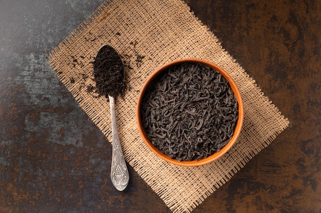 Foglie di tè nere secche in una ciotola di argilla e un cucchiaio vintage su sfondo scuro vista dall'alto Tè nero a foglia larga e piccola