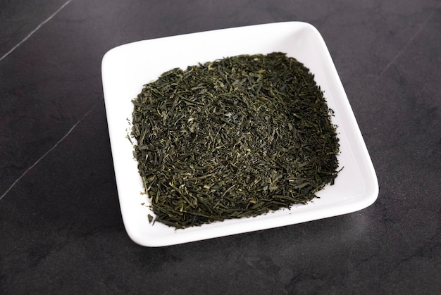 Foglie di tè verde asciutte in un piatto quadrato bianco su sfondo nero