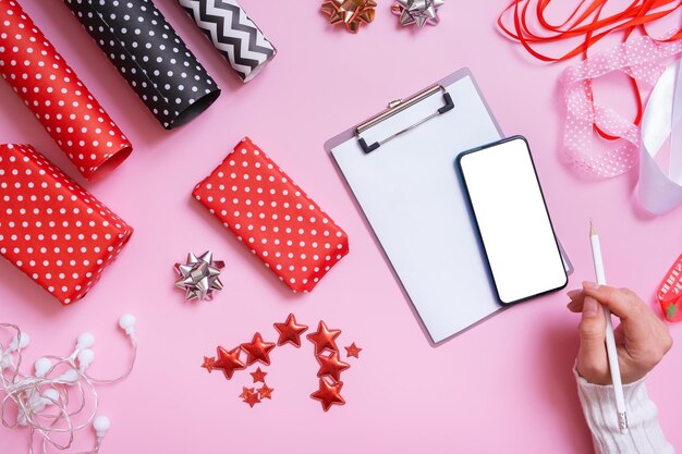 Foglio bianco di telefono di carta e mani femminili che avvolgono i regali di natale distesi su sfondo rosa