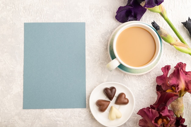 foglio di carta blu con tazza di cioffee, viola cioccolato e fiori di iris bordeaux su grigio.