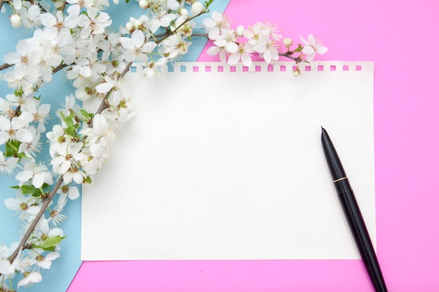Foglio vuoto di blocco note con penna su uno sfondo rosa-blu con fiori in fiore. Copia spazio per il testo di primavera.