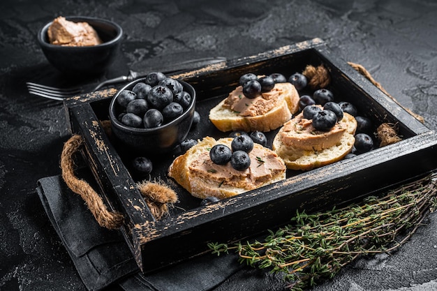 Foie gras toasts, duck liver pate and fresh blueberry in wooden tray. Black background. Top view.