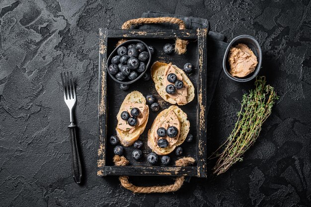 Foie gras toasts, duck liver pate and fresh blueberry in wooden tray. Black background. Top view.