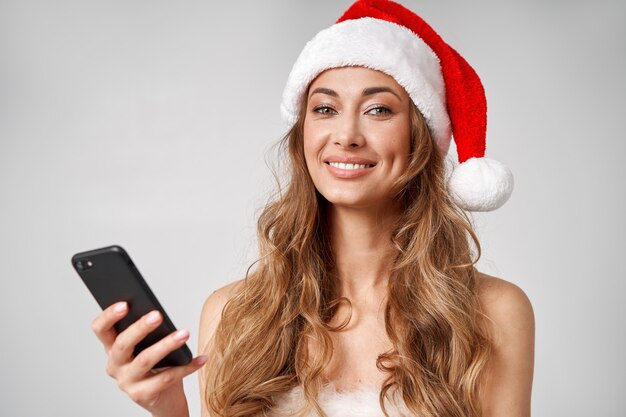 Fondo bianco dello studio del cappello di Santa di natale della donna con lo smartphone a disposizione