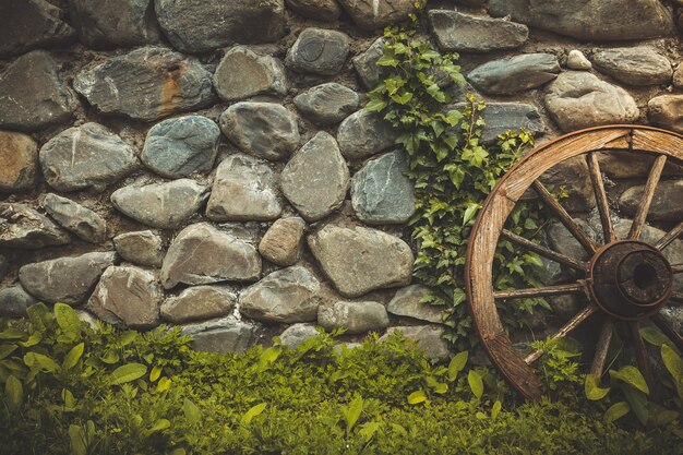 Fondo di struttura della parete di pietra. Il modello del vecchio muro di pietra con la ruota antica e la crescita dell'erba verde. Filtro tonificante vintage retrò