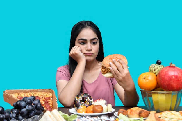 foodie ragazza seduta al tavolo della frutta sorridente tenendo hamburger con la bocca aperta indiano modello pakistano