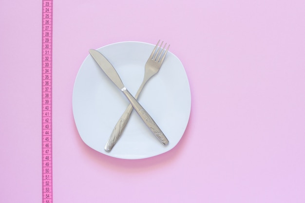 Forcella e coltello attraversati sul piatto e sulla misura di nastro bianchi su fondo rosa.