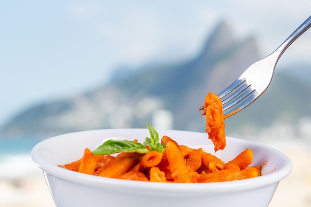 Forchetta con penne in salsa di pomodoro in una ciotola bianca con la spiaggia di ipanema a rio de janeiro sullo sfondo