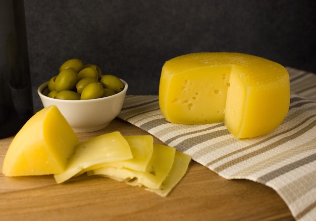 Forma di formaggio con un pezzo tagliato su una tavola di legno. Olive verdi e pezzi di formaggio nelle vicinanze.