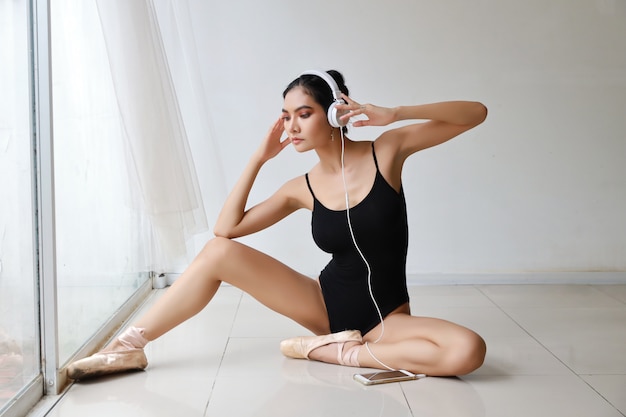 Forma sbalorditiva della donna asiatica affascinante che indossa abiti sportivi neri che si siedono con la cuffia. Musica d'ascolto della bella donna dal telefono cellulare mentre preparando balletto