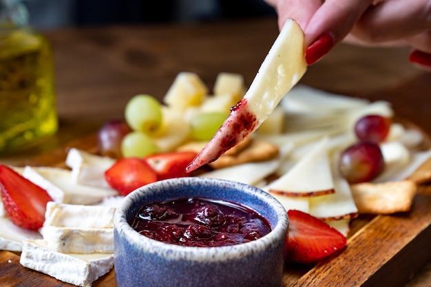 Formaggi assortiti con frutta e marmellata su tavola di legno. le mani delle donne intingono un pezzo di formaggio nella marmellata. servire la tavola e gli spuntini al vino per il banchetto.
