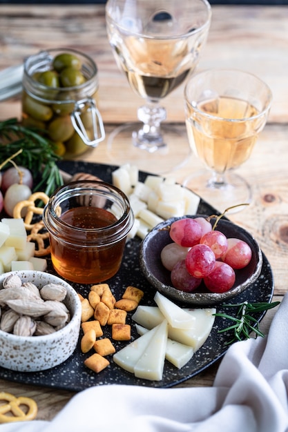 Formaggi assortiti su un piatto scuro su un tavolo di legno. Formaggio, uva, noci, olive, rosmarino e un bicchiere di vino bianco.