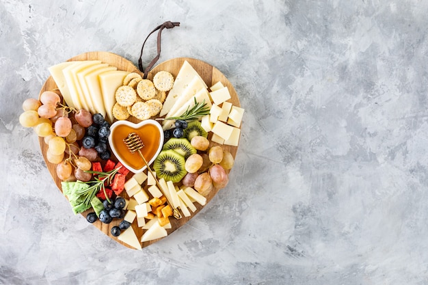 Formaggi assortiti su un tagliere di legno a forma di cuore. Formaggio, uva, noci, olive, rosmarino e un bicchiere di vino bianco. vista dall'alto, copia dello spazio